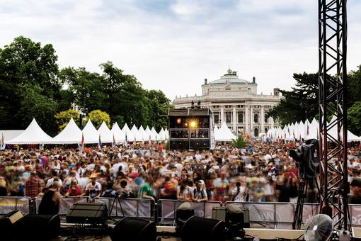 Das Foto zeigt den Wiener Rathausplatz, von einer Bühne aus auf das Burgtheater gerichtet, vor der Bühne eine Menschenmenge, am Rand weiße Zelte mit Spitzdach